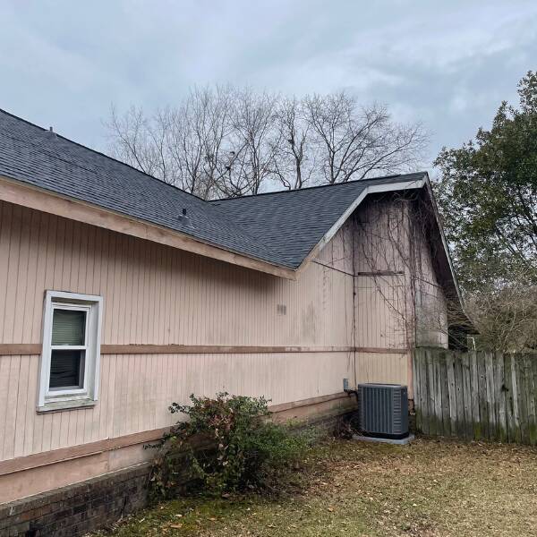 Side view of a beige house with a window, an outdoor AC unit, and a wooden fence under a cloudy sky.